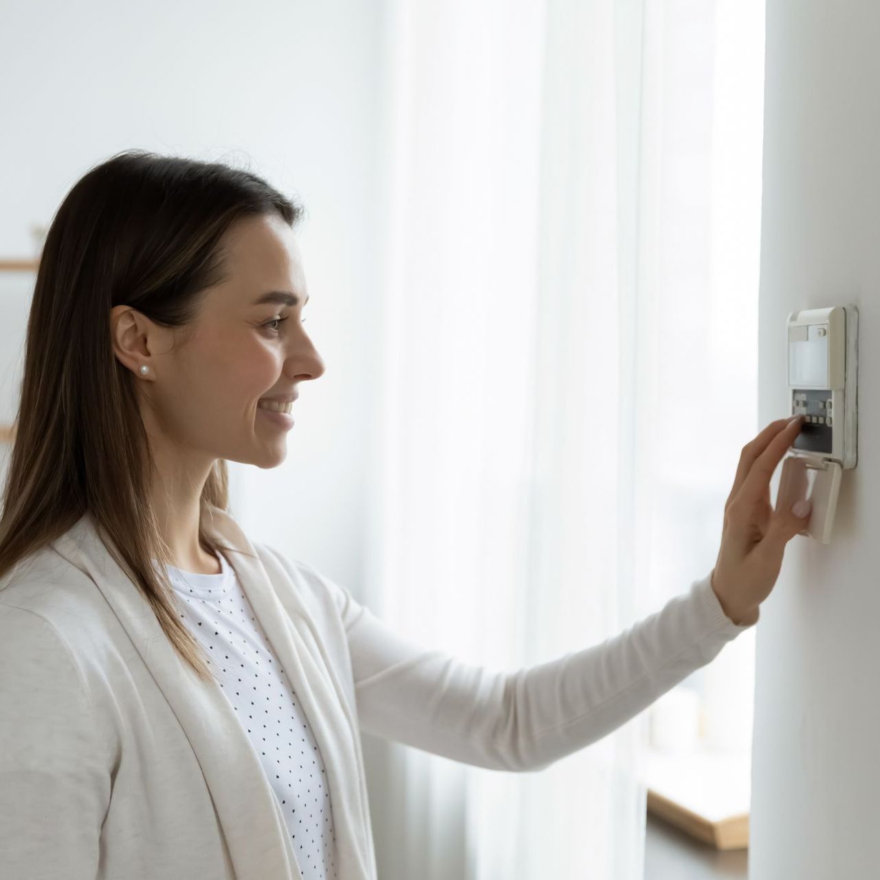 Woman adjusting a thermostat on a white wall; interior setting.