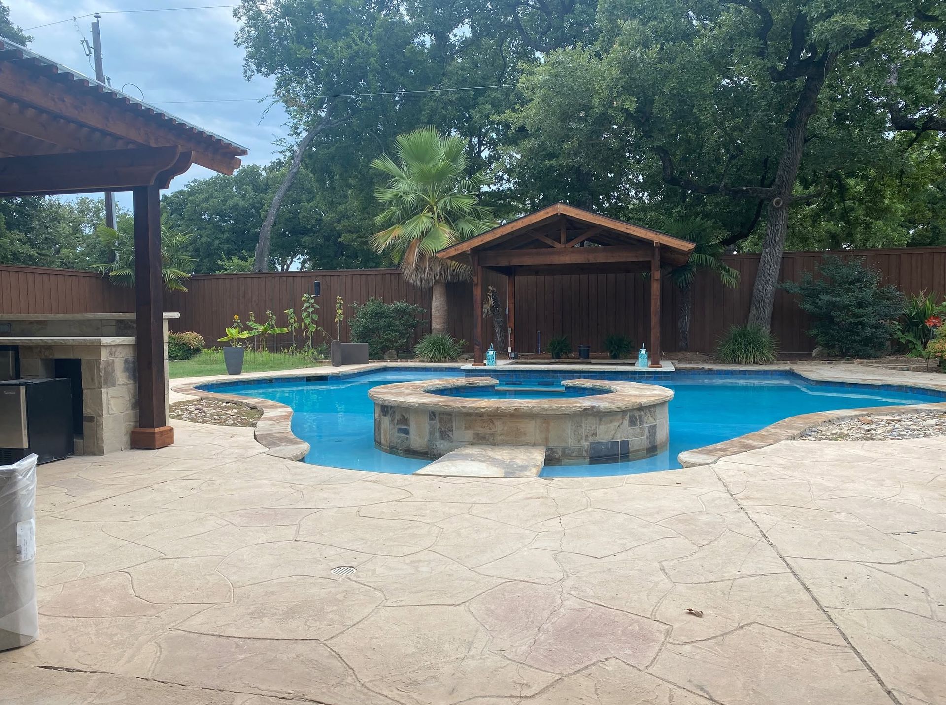 Backyard pool with stone hot tub, covered patio, and wooden cabana under trees