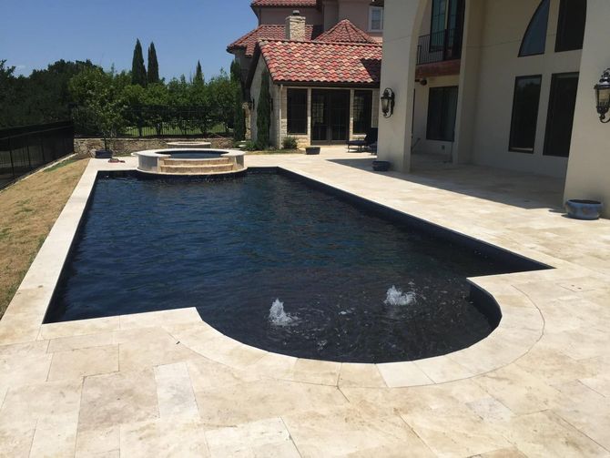 Rectangular backyard swimming pool beside a stone patio and house, with a small fountain spray at one end