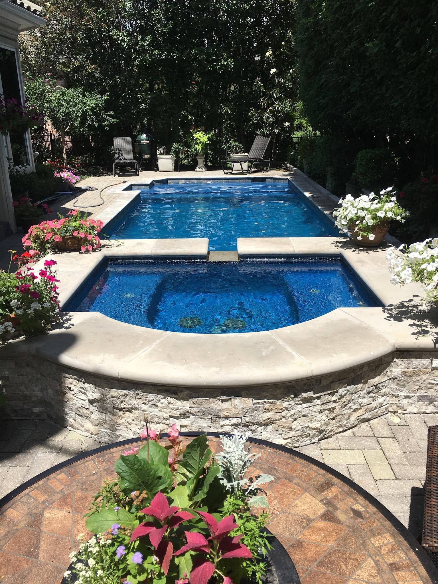 Backyard pool with a small spa, blue water, stone deck, and flowering plants, framed by trees