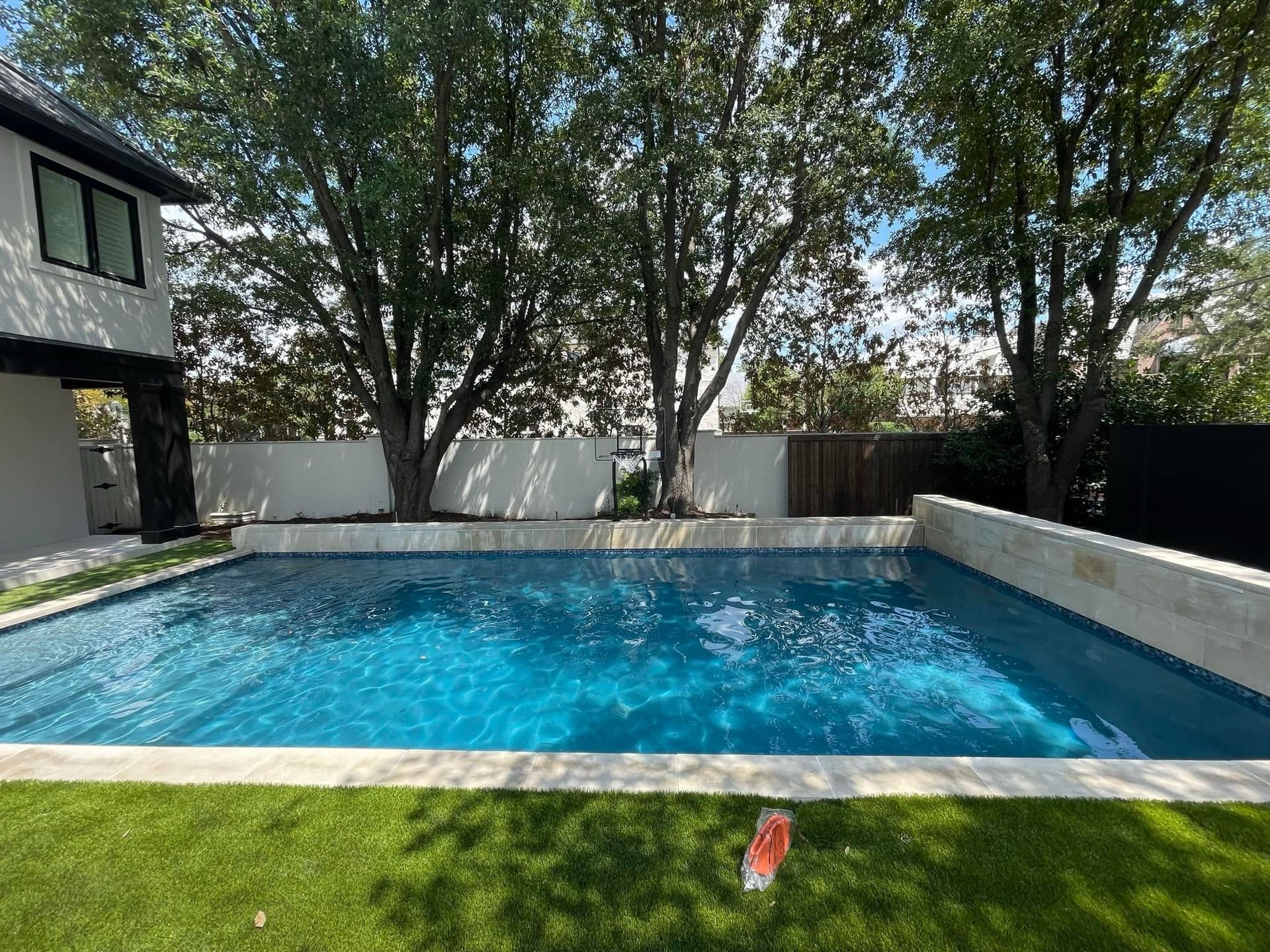 Backyard swimming pool with turquoise water, surrounded by trees, a patio, and a house on the left