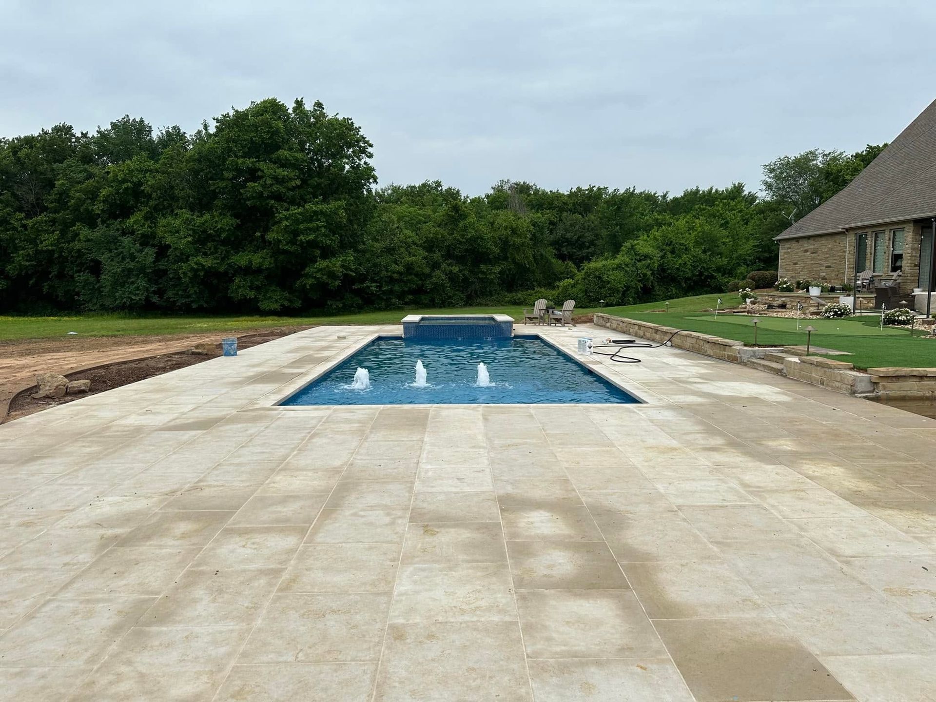 Backyard patio with a rectangular swimming pool, stone deck, and trees beyond