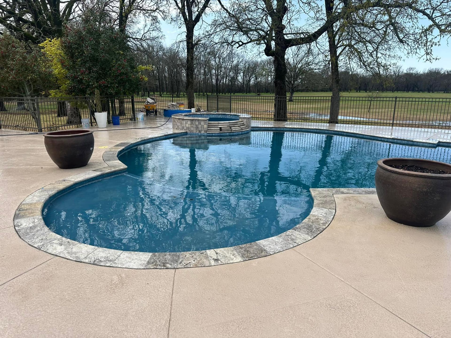 Backyard pool with blue water, stone coping, and large planters beside a fenced grassy yard