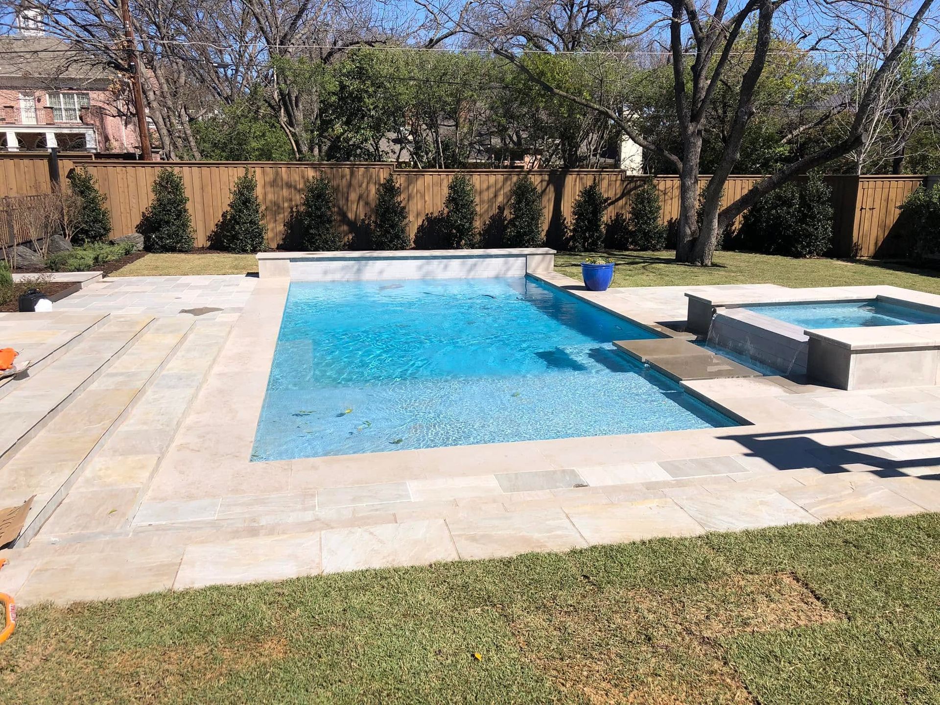 Backyard swimming pool with a stone deck, grass, and trees along the fence line.