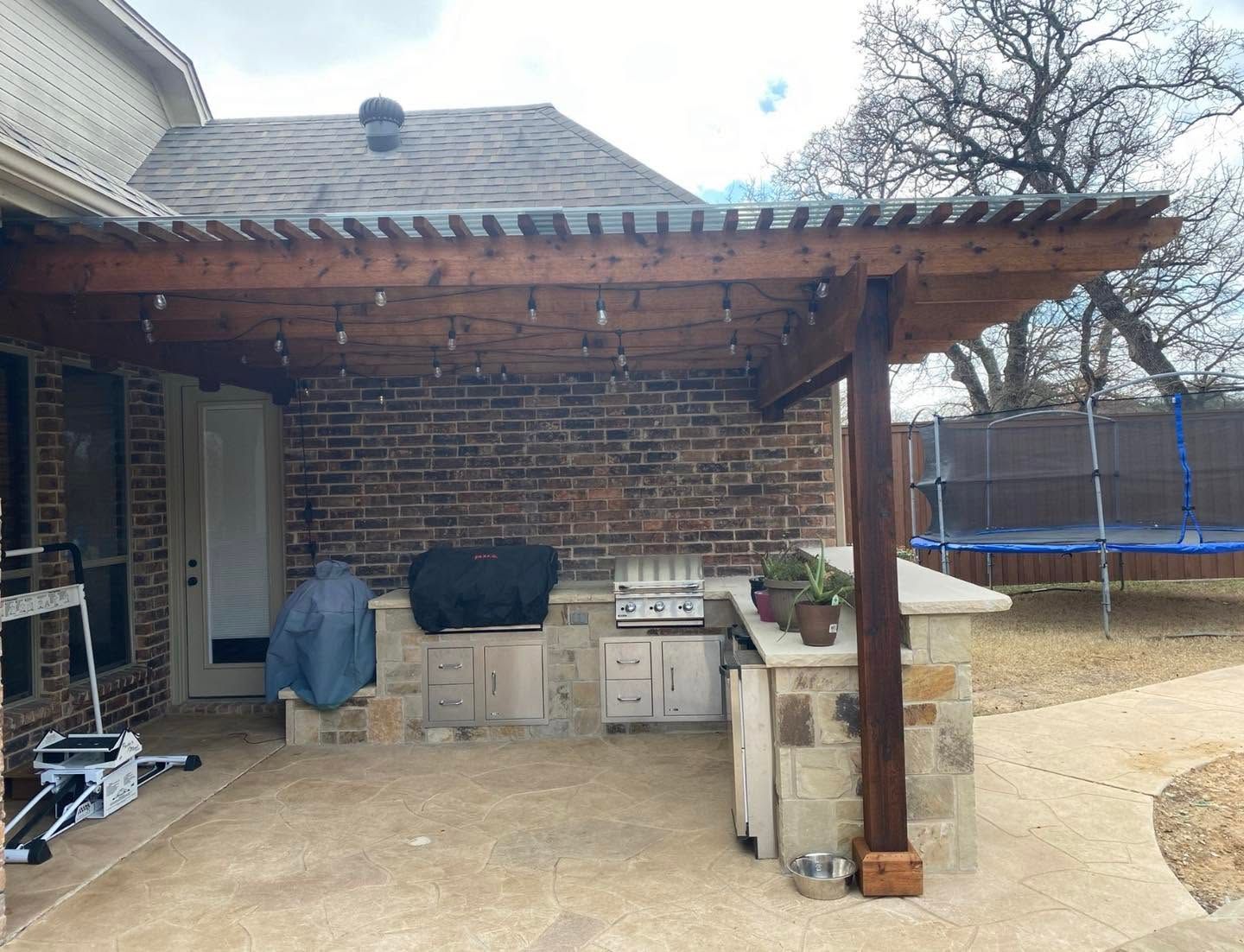Brick backyard patio with a covered pergola, grill, and trampoline in the background