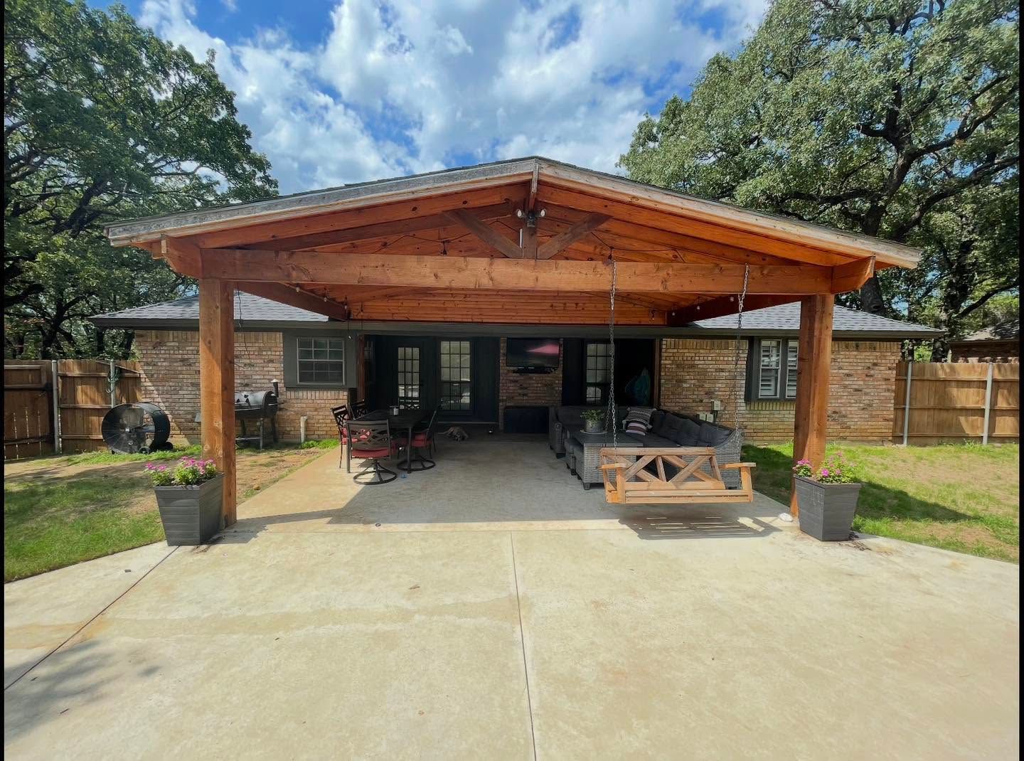 Wooden covered patio with concrete driveway, outdoor seating, and green yard under a partly cloudy sky