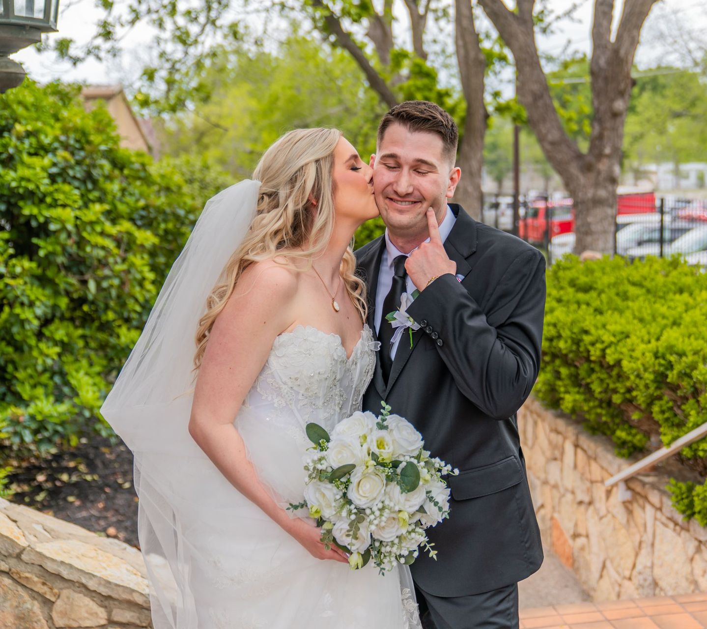 Bride and groom in wedding attire smiling outdoors near stone steps and trees