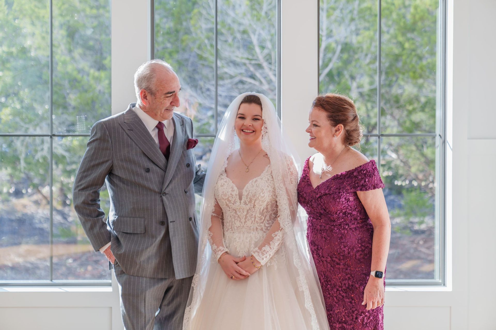 Three people posing by large windows, with a bride in a white dress between two formally dressed companions.