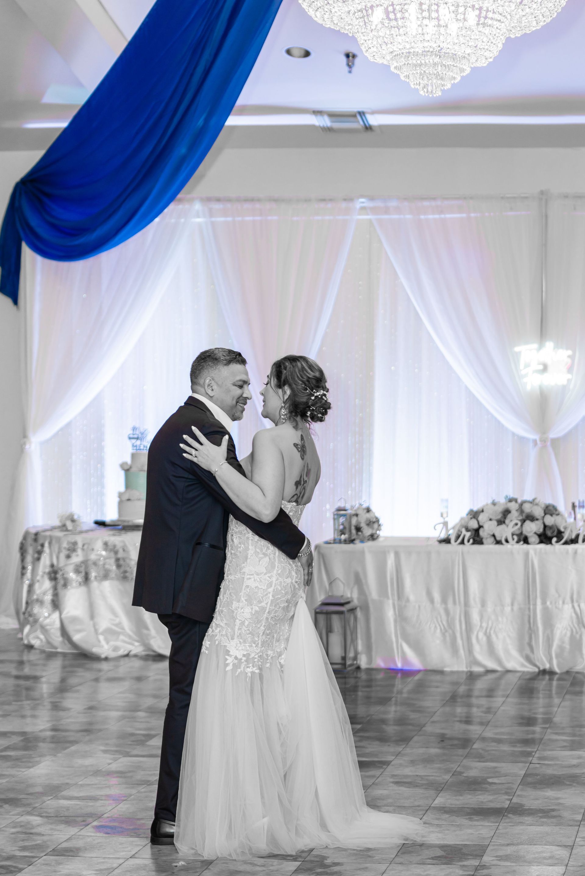 Couple dancing at a wedding reception in a decorated ballroom, with blue and white drapes.