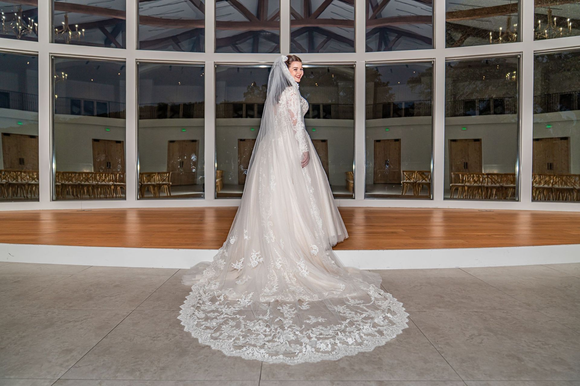 Bride in a white lace wedding gown standing in a glass atrium with a long train.