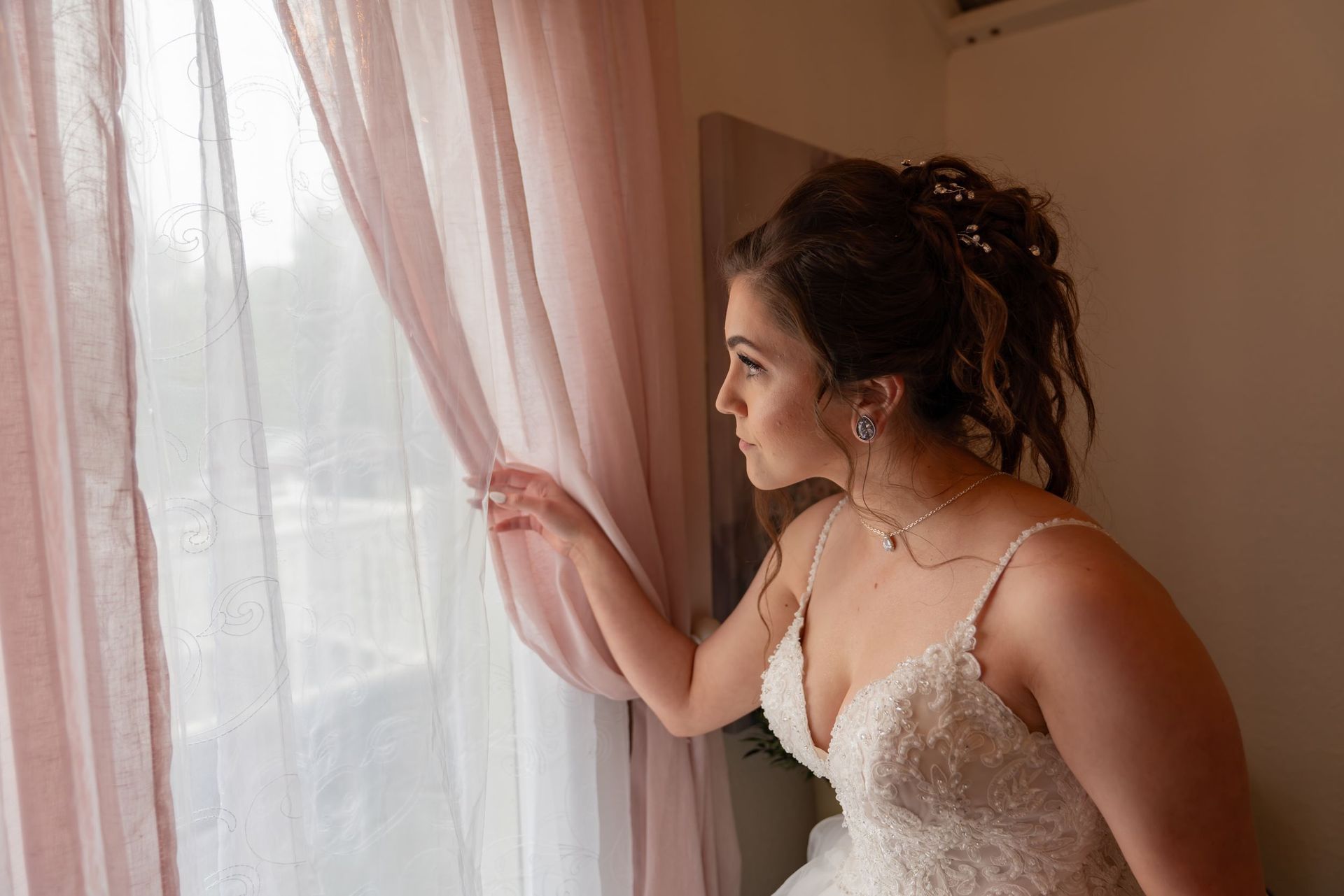 Woman in a white lace dress gently parts sheer curtains by a sunlit window.