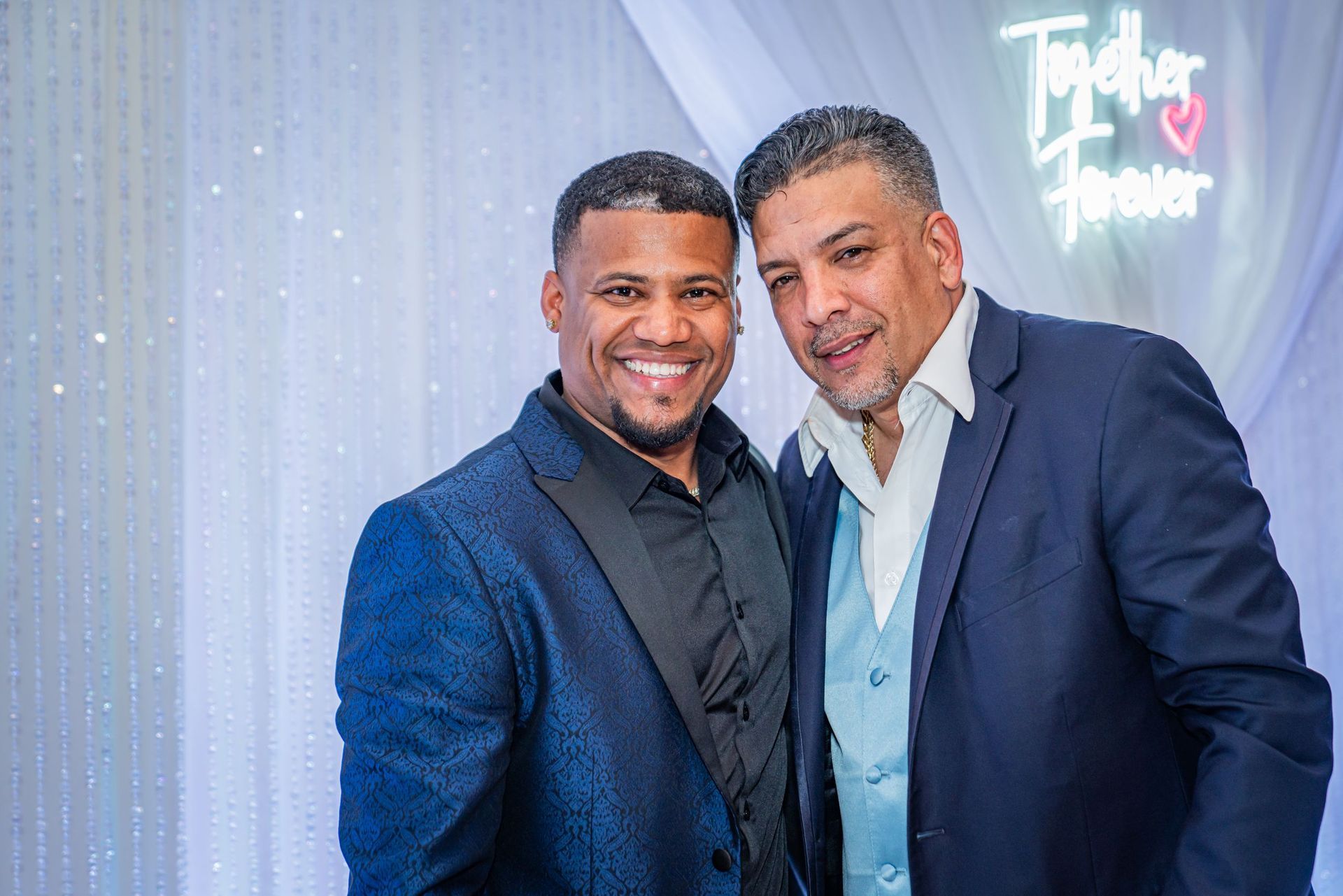 Two men in blue suits smiling and posing at an event with a glowing neon sign background