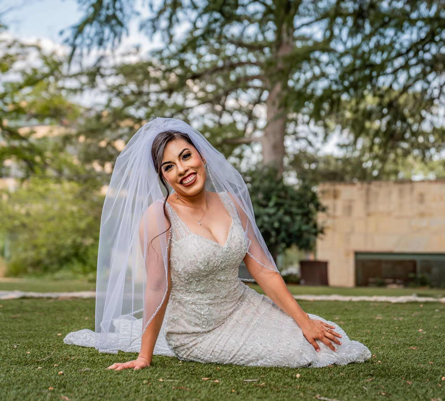 Bride in a white wedding dress and veil sitting on grass in a park, smiling at the camera.