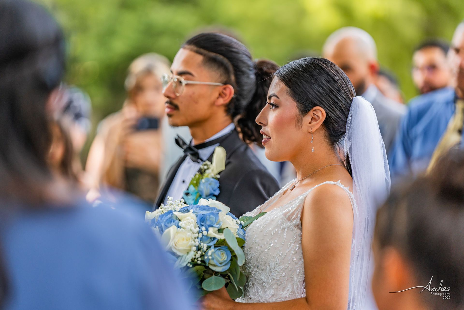 Bride in white gown and groom in gray suit walking outdoors, with guests blurred in the background.