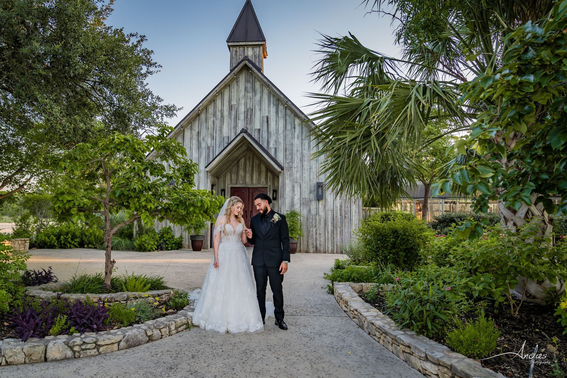 Bride and groom posing on a garden path in front of a rustic chapel