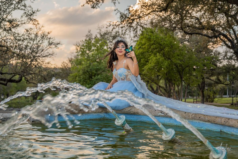 Woman in a blue dress sitting by a pond with water splashing around her at sunset