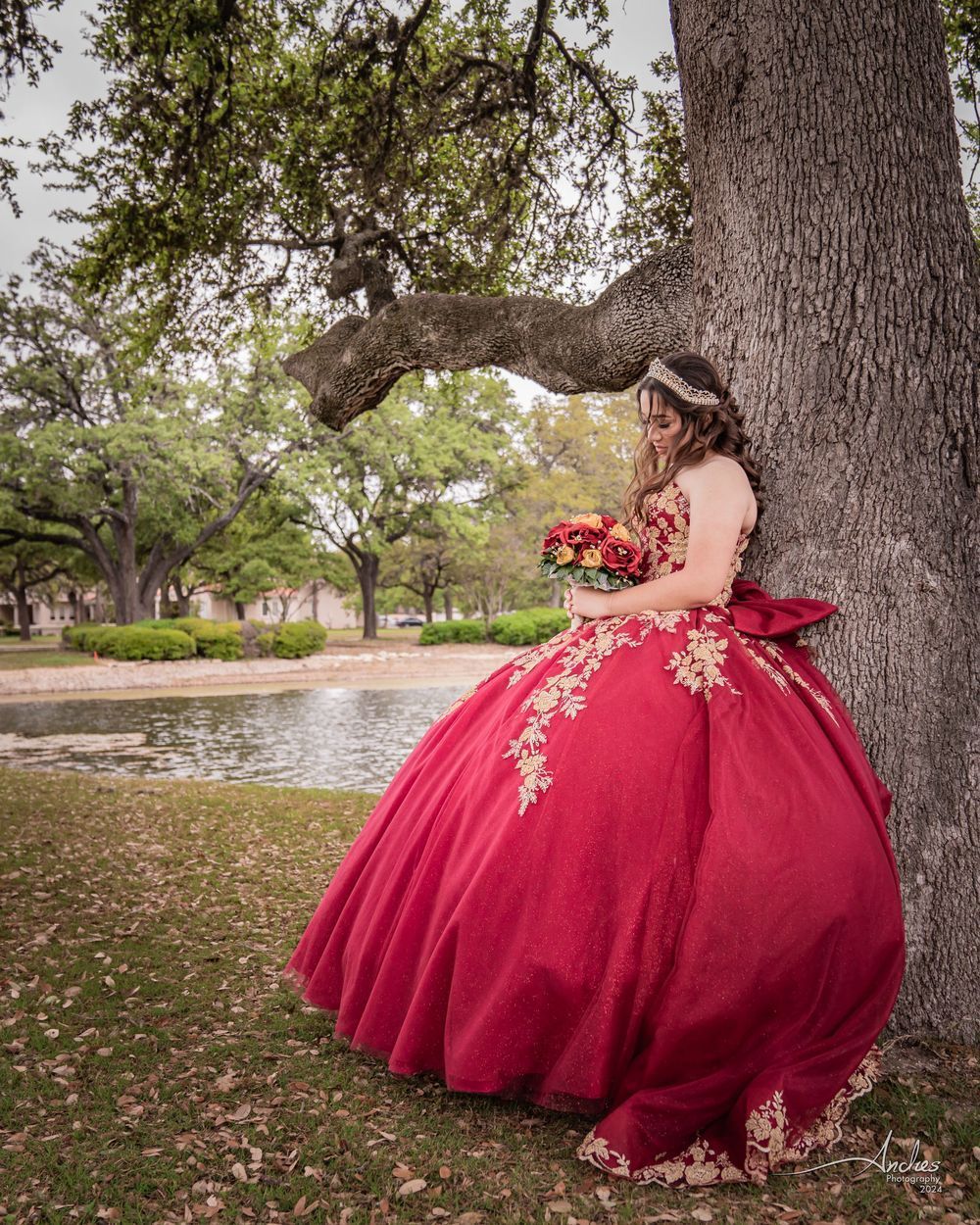 Woman in a bright red ball gown holding a bouquet beside a large tree by a pond