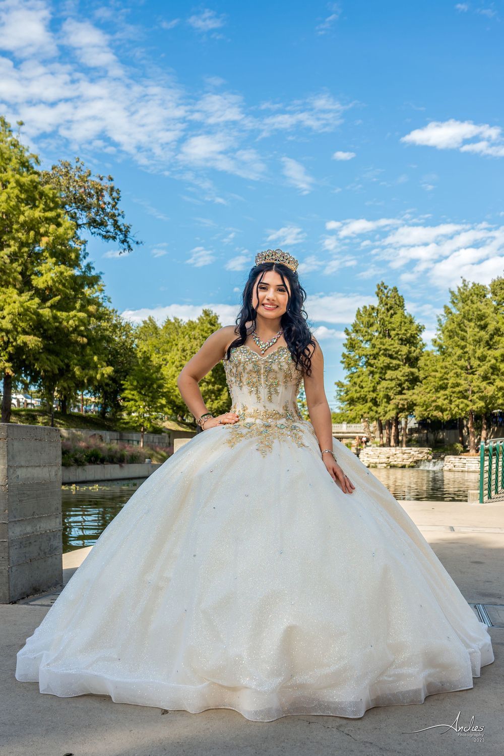 Woman in a white gown standing outdoors on a tree-lined path under a blue sky