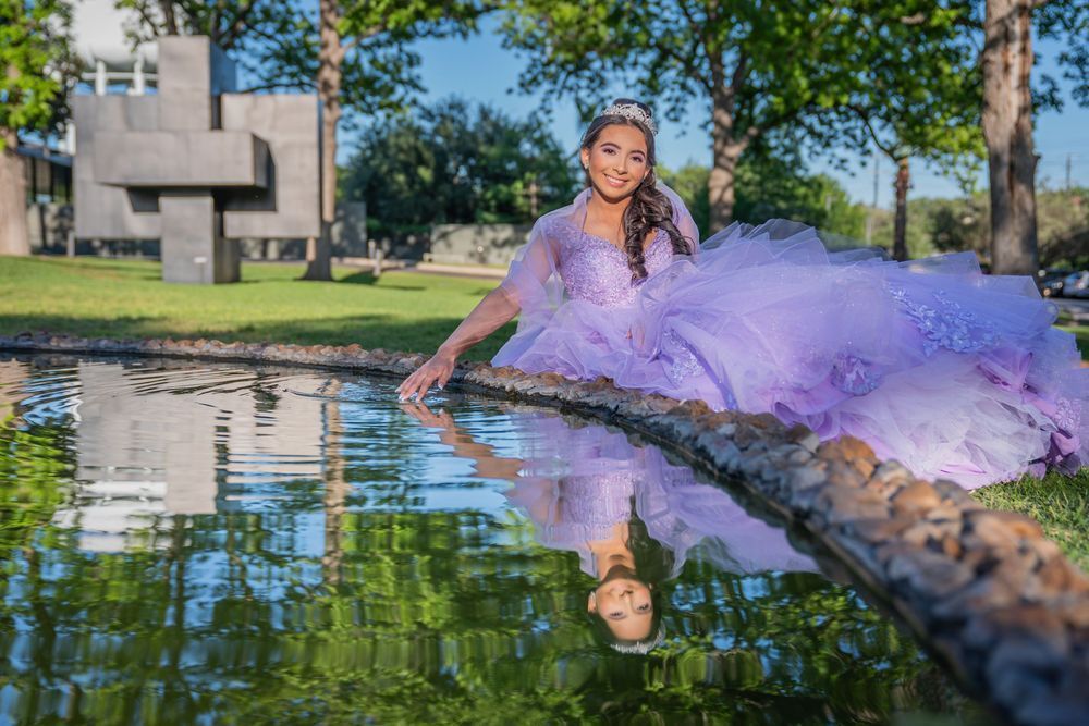 Woman in a lavender gown seated by a reflective pond in a park, with a stone structure and trees behind her