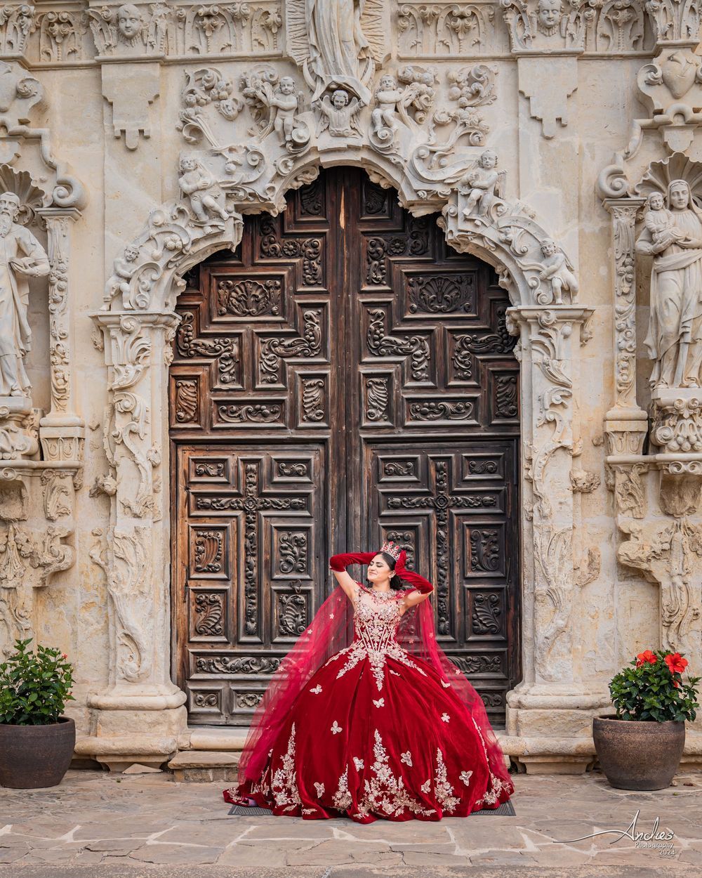 Woman in a red gown standing before an ornate carved doorway with stone columns and potted flowers