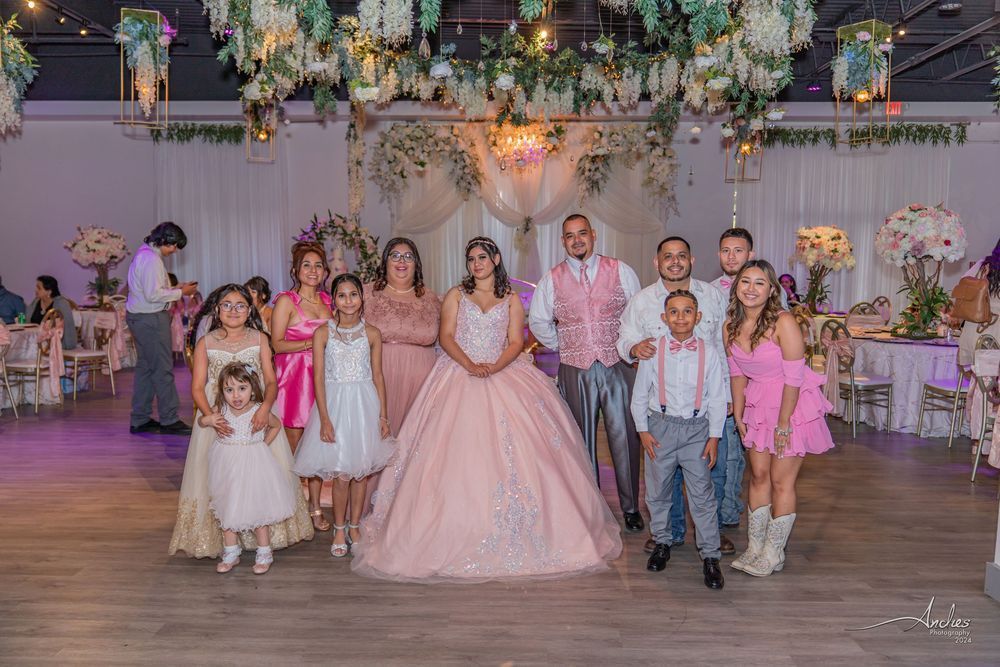Group posing at a pink-themed celebration in a decorated ballroom, with flowers and string lights overhead