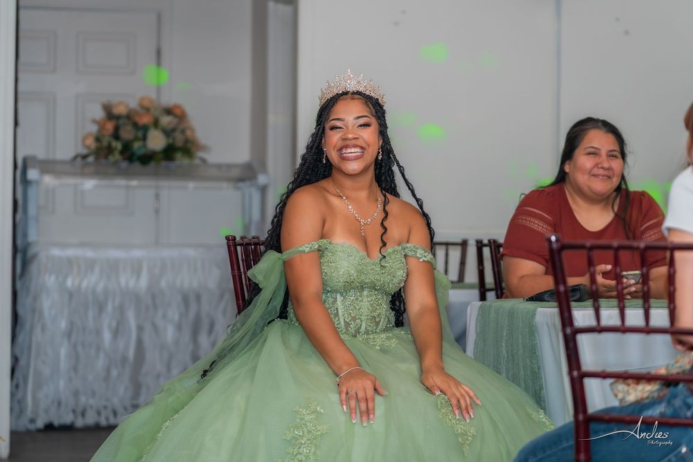 Woman in a green gown seated at a decorated indoor event, smiling beside a table of flowers.