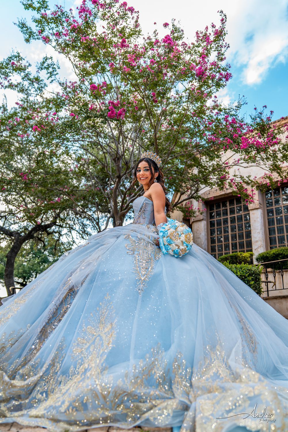 Woman in a light blue gown holding a bouquet, standing in a garden with blooming pink flowers and a building behind her.