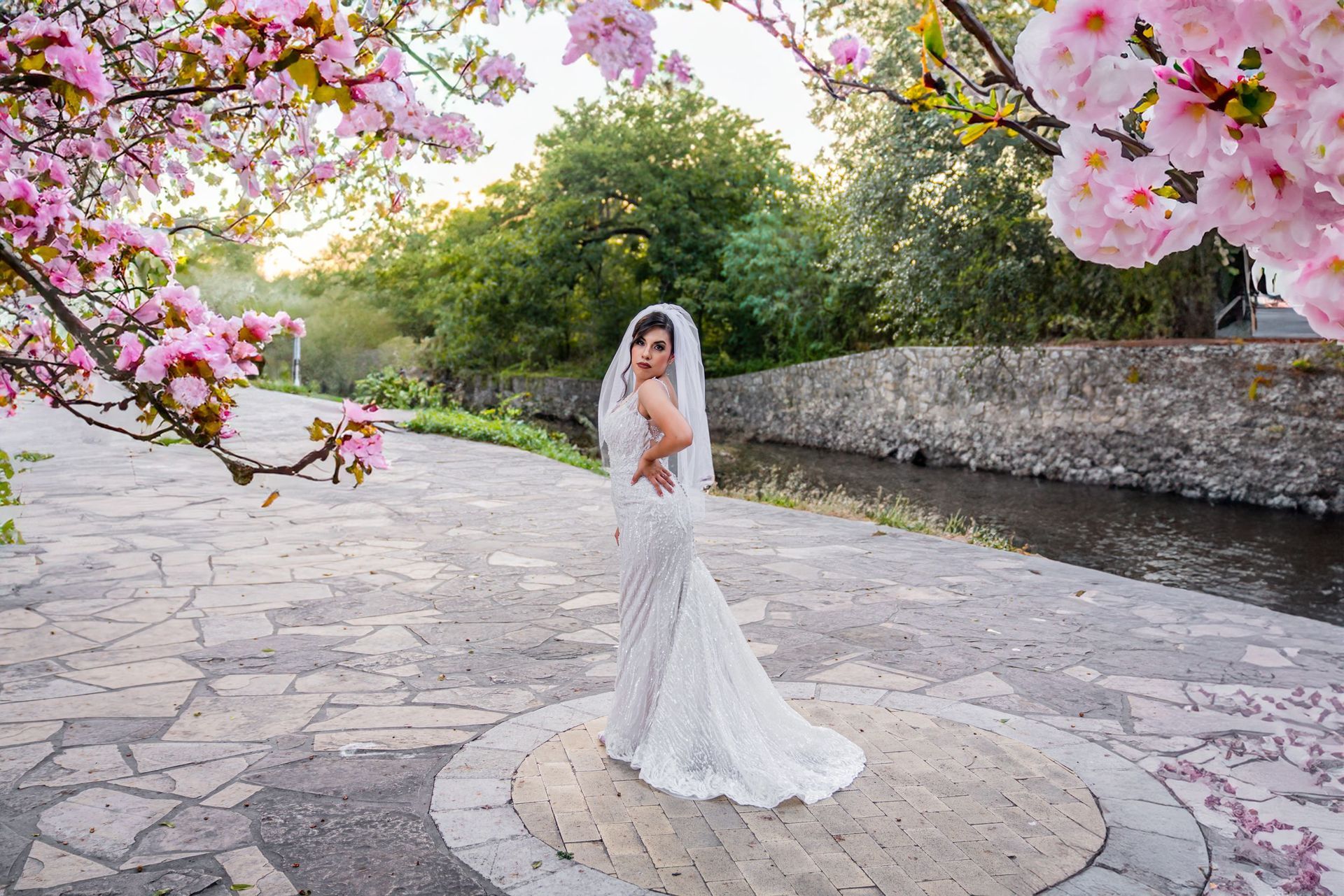 Bride in a white wedding dress standing under pink cherry blossoms beside a stone path and stream.
