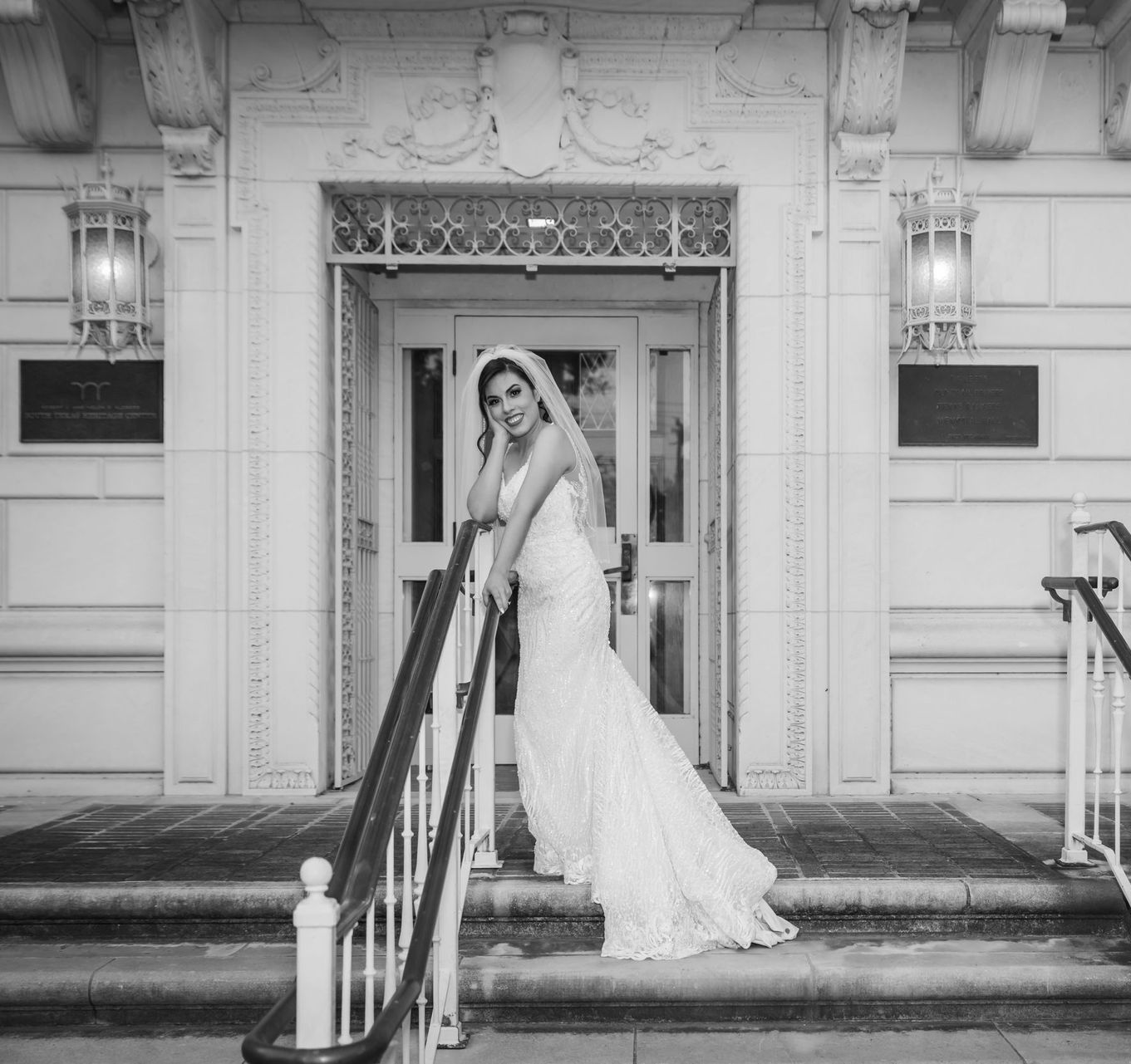 Bride in a white gown standing on steps outside a building, holding a bouquet under an ornate doorway