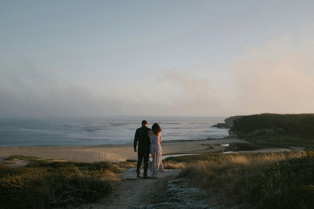 Couple standing on a coastal path at sunset overlooking a calm beach and ocean