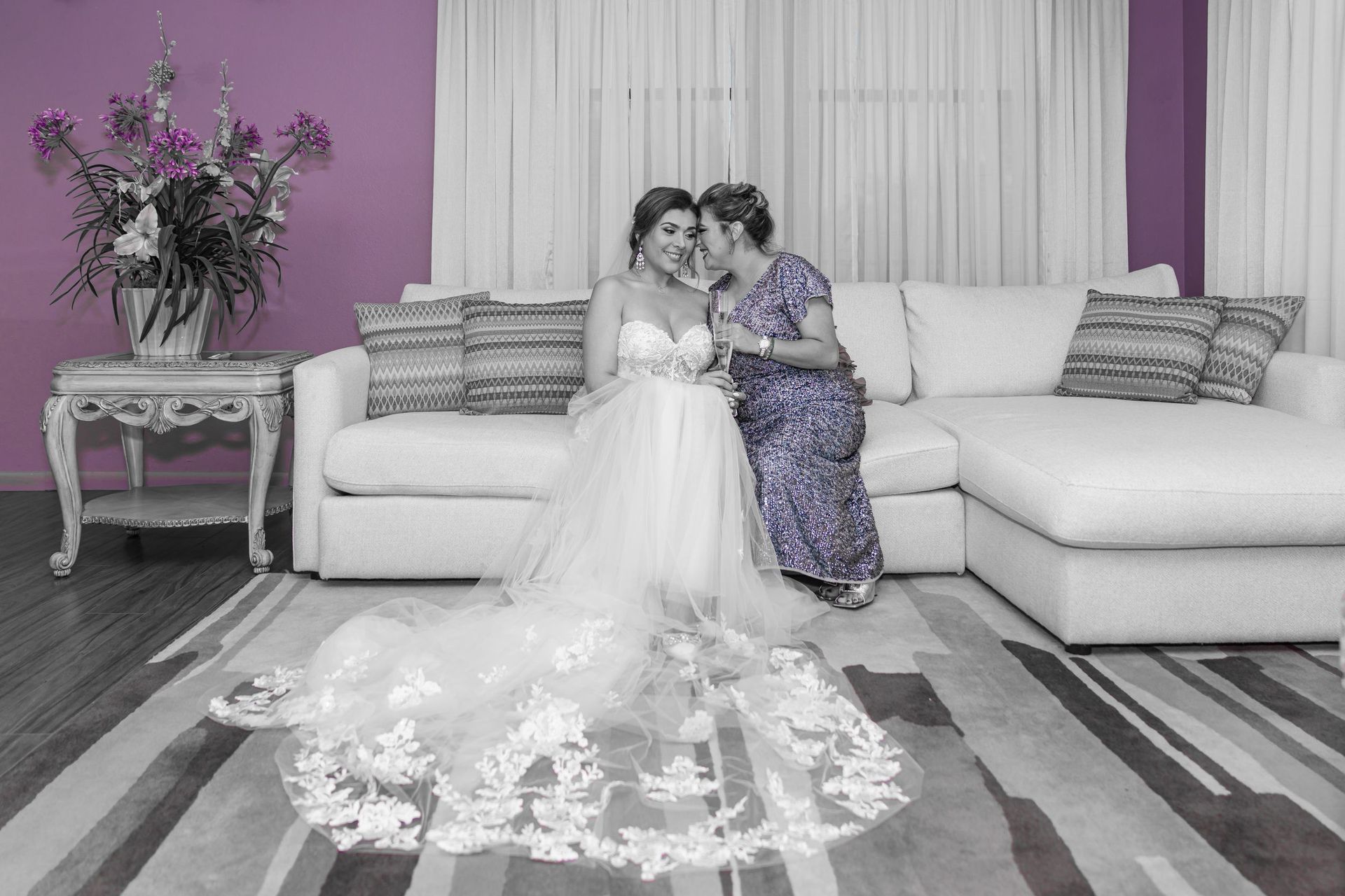 Two women in wedding dresses sitting together on a white sectional in a living room, one with a long train.