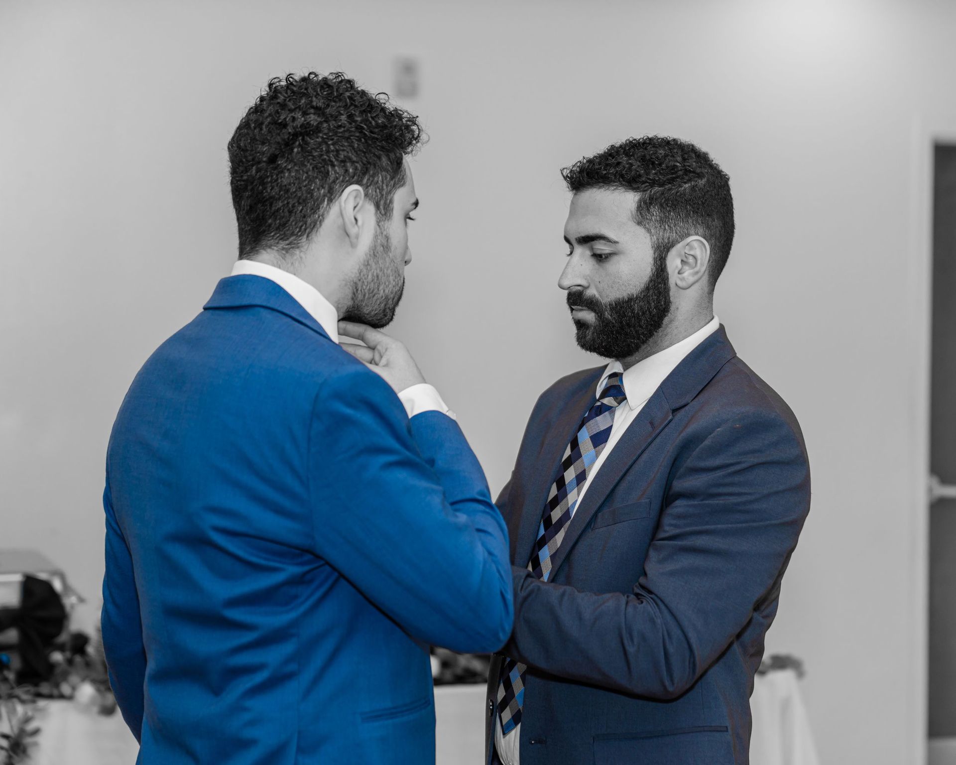Two men in blue suits face each other, adjusting a tie in an office setting.
