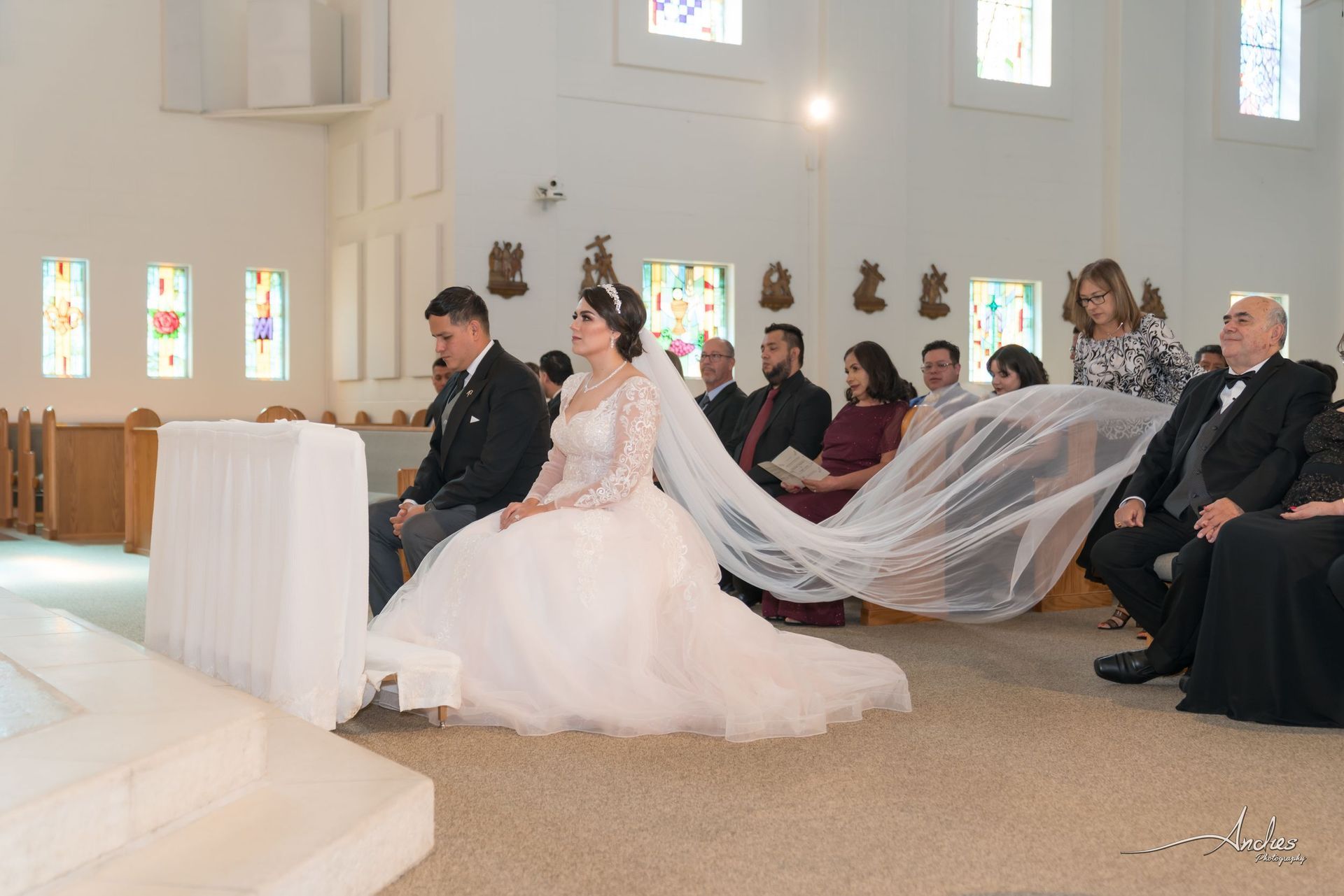 Wedding ceremony in a bright church, with bride and groom seated before guests and her veil flowing back.