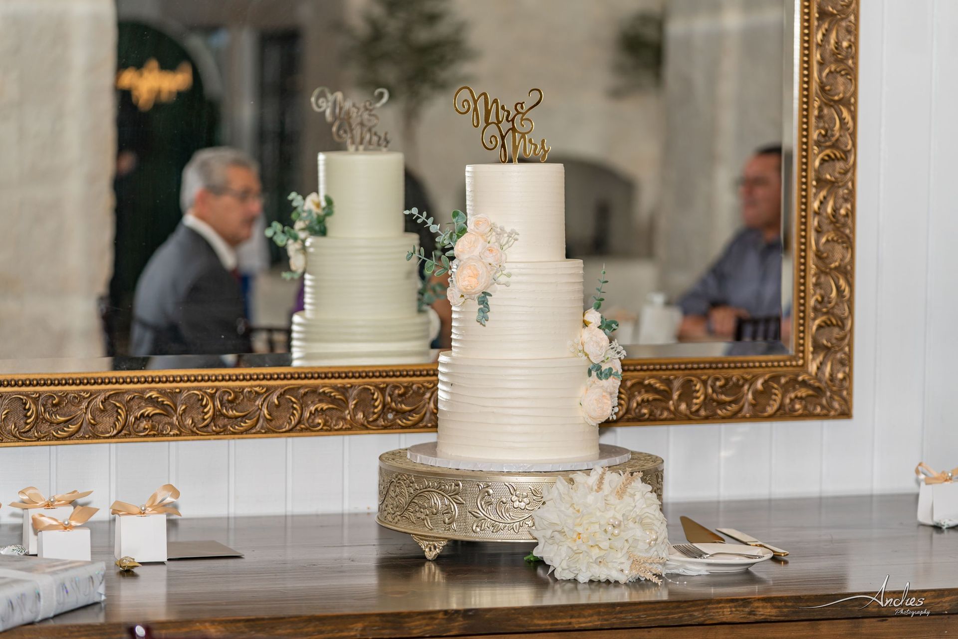 White tiered cake with gold topper and flowers on a decorated table in front of a mirror