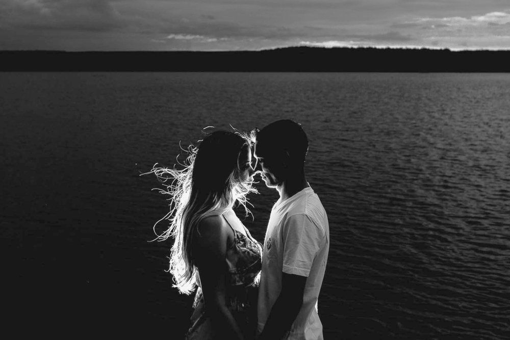 Silhouetted couple facing each other by a lake at sunset, with bright backlighting and rippling water.