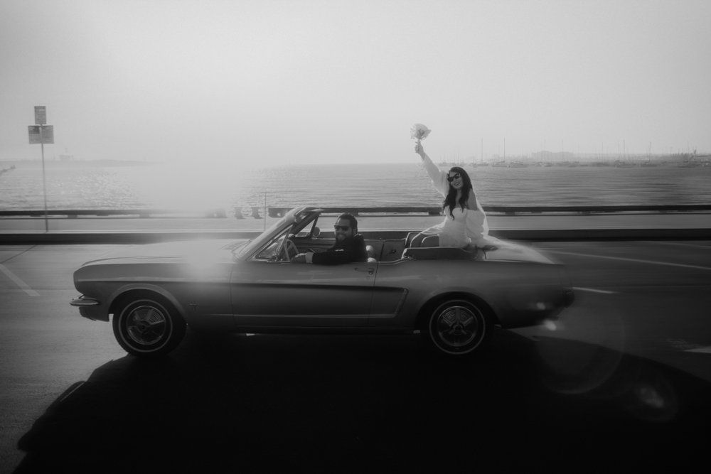 Woman standing through car sunroof beside a vintage car on a foggy road by the waterfront
