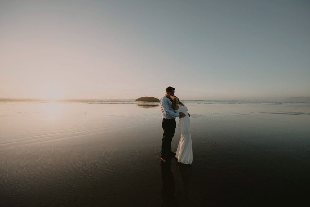Couple embracing on a calm beach at sunset, with soft reflections on the water.