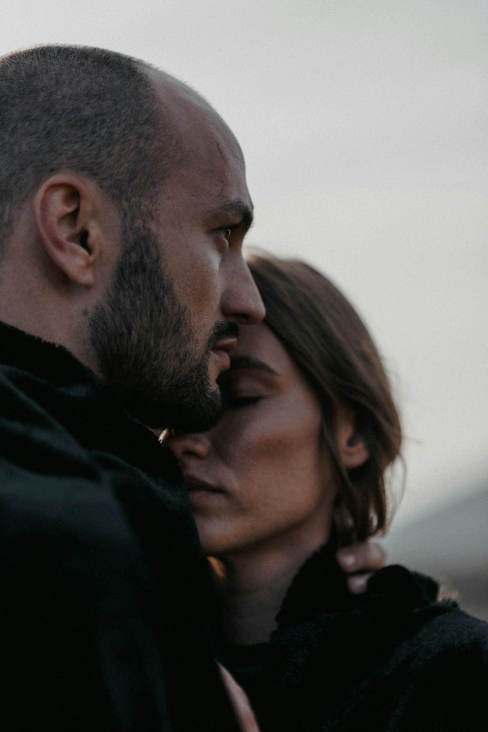 Two people in dark clothing embrace closely outdoors at dusk, with a soft, blurred background.