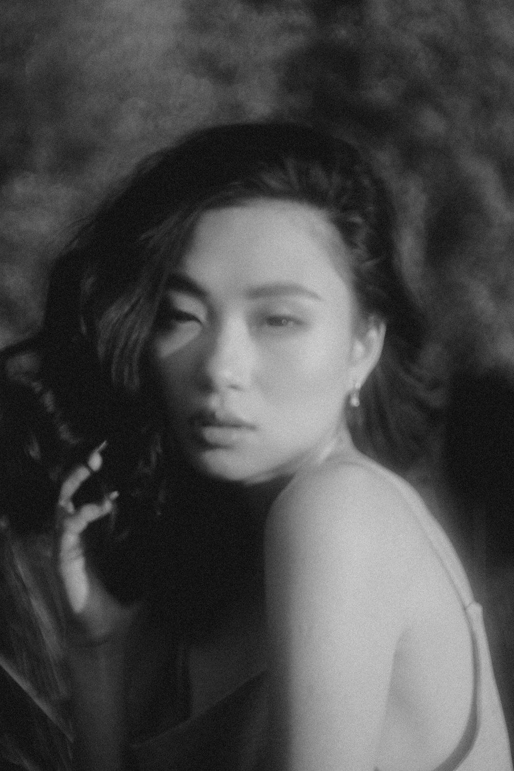 Black-and-white portrait of a person with long hair, looking over their shoulder against a dark, blurred background