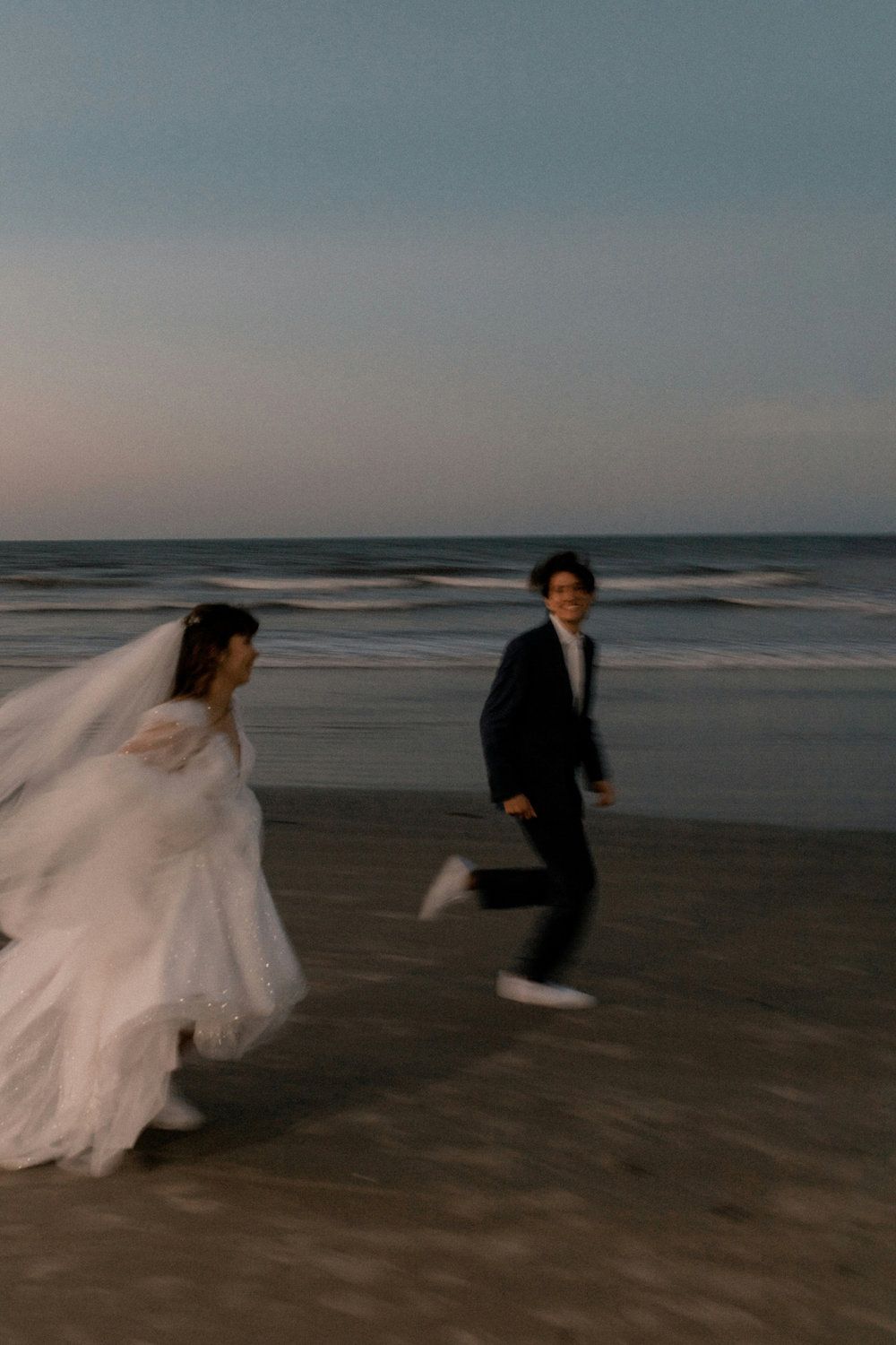 A bride and groom run along a beach at dusk, with the bride’s veil flowing behind them.