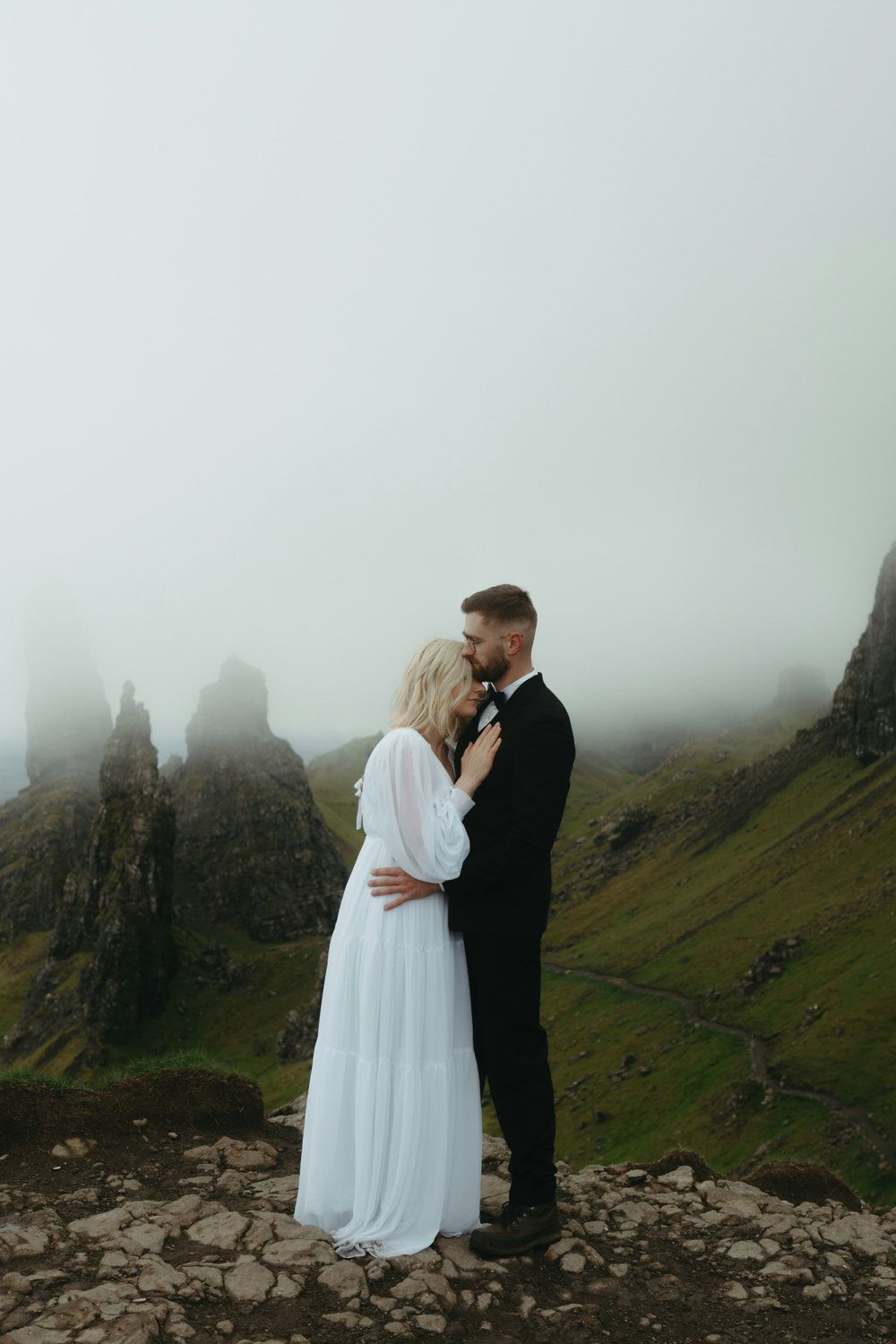 Couple embracing on a misty cliff, with rocky peaks and green hills in the background