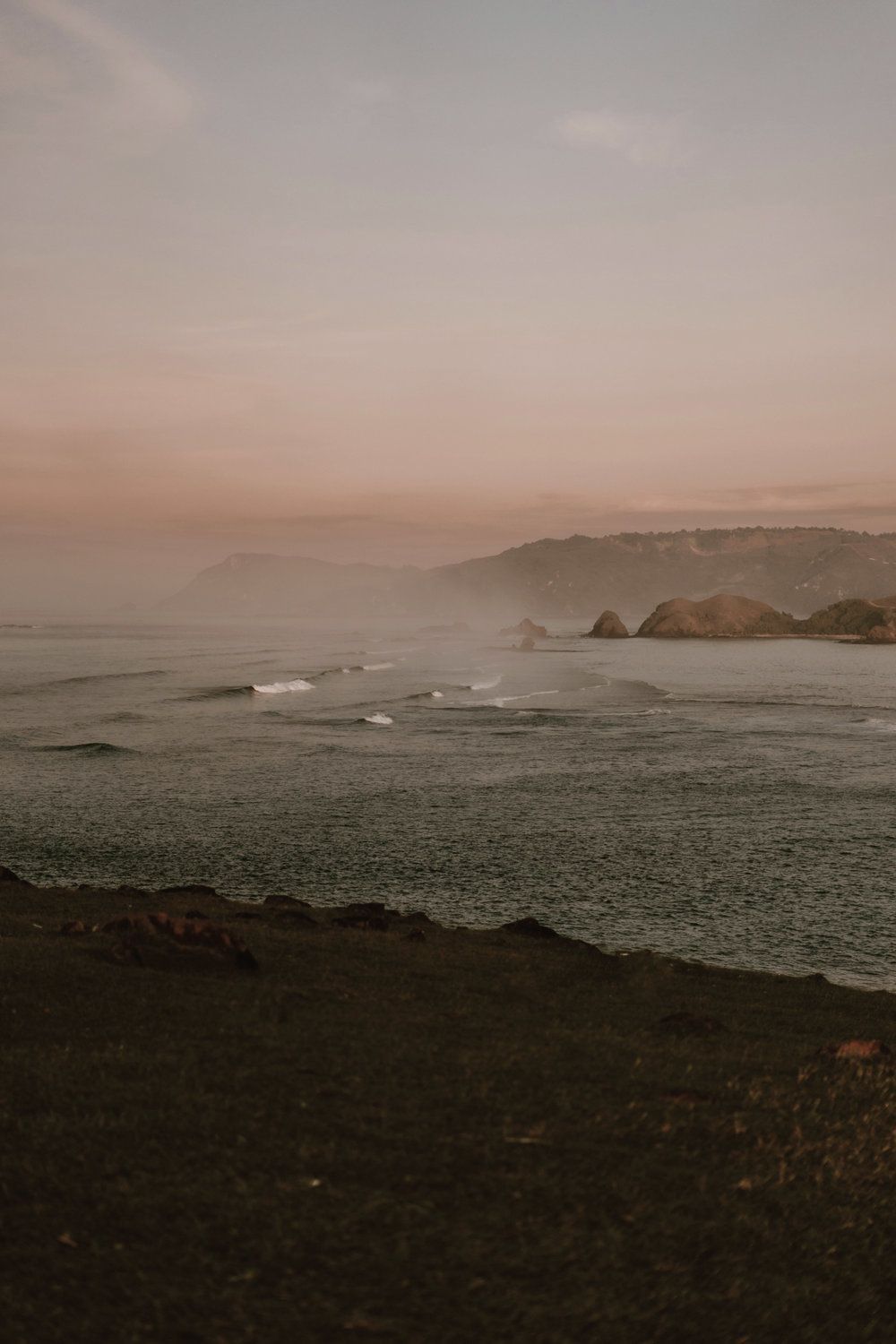 Misty shoreline at dusk with dark sandy beach, calm waves, and distant hills under a pink sky