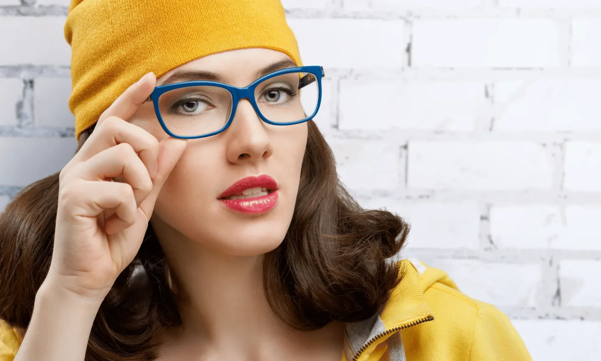 A woman wearing glasses and a yellow hat is standing in front of a white brick wall.