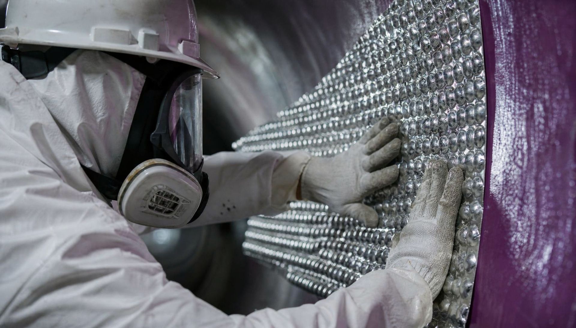 Trabalhador com equipamento de proteção individual aplicando isolamento prateado em um painel de parede roxo.