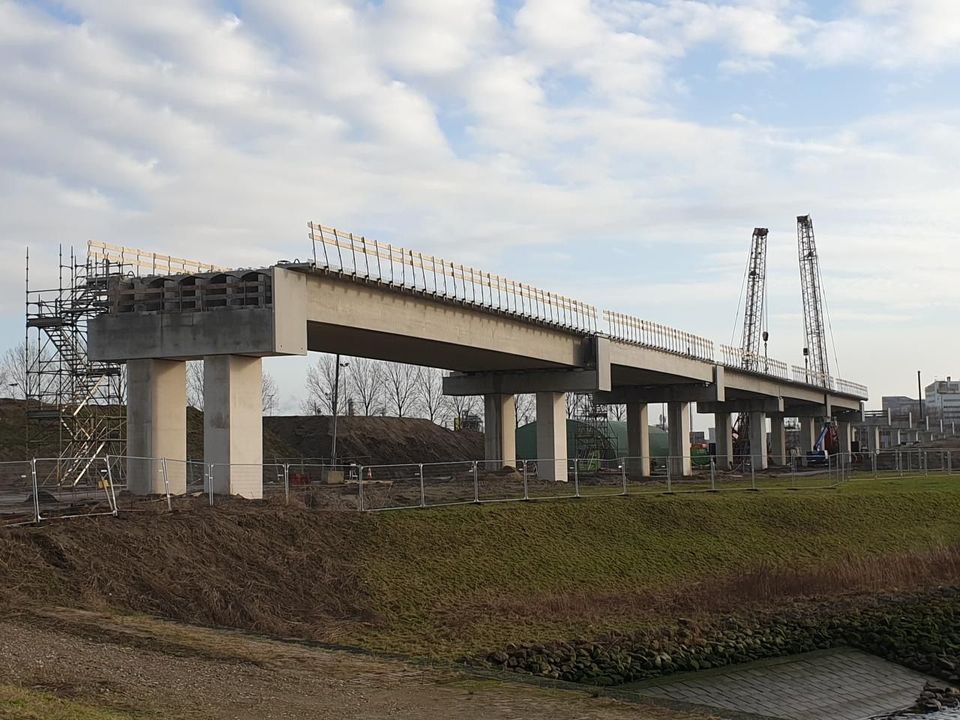 Een verhoogde betonnen brug in aanbouw over een braakliggend veld met struiken tegen een bewolkte hemel.