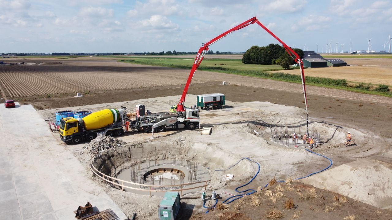 Een betonpomp stort cement in een ronde funderingsput op een bouwterrein. Een gele cementmixer is aanwezig.