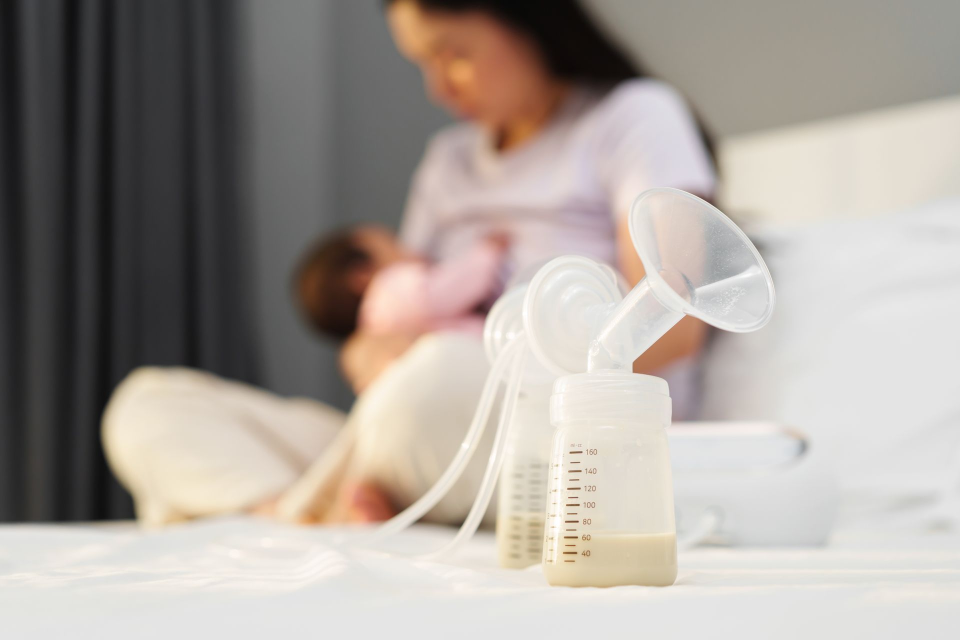 A woman is breastfeeding her baby on a bed while a bottle of milk is on the table.