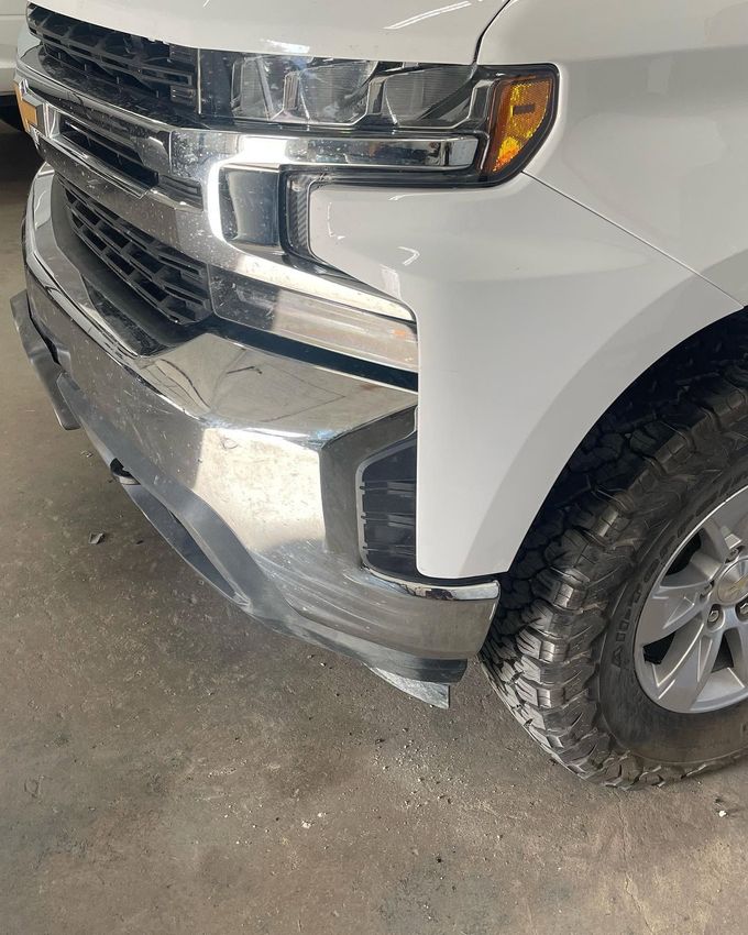 Damaged front end of a white pickup truck in a garage, with dented bumper and grille.