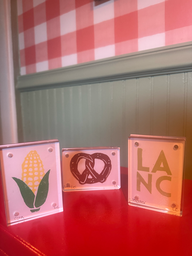 Three small metal signs on a red table: corn, pretzel, and “LA NC” designs.