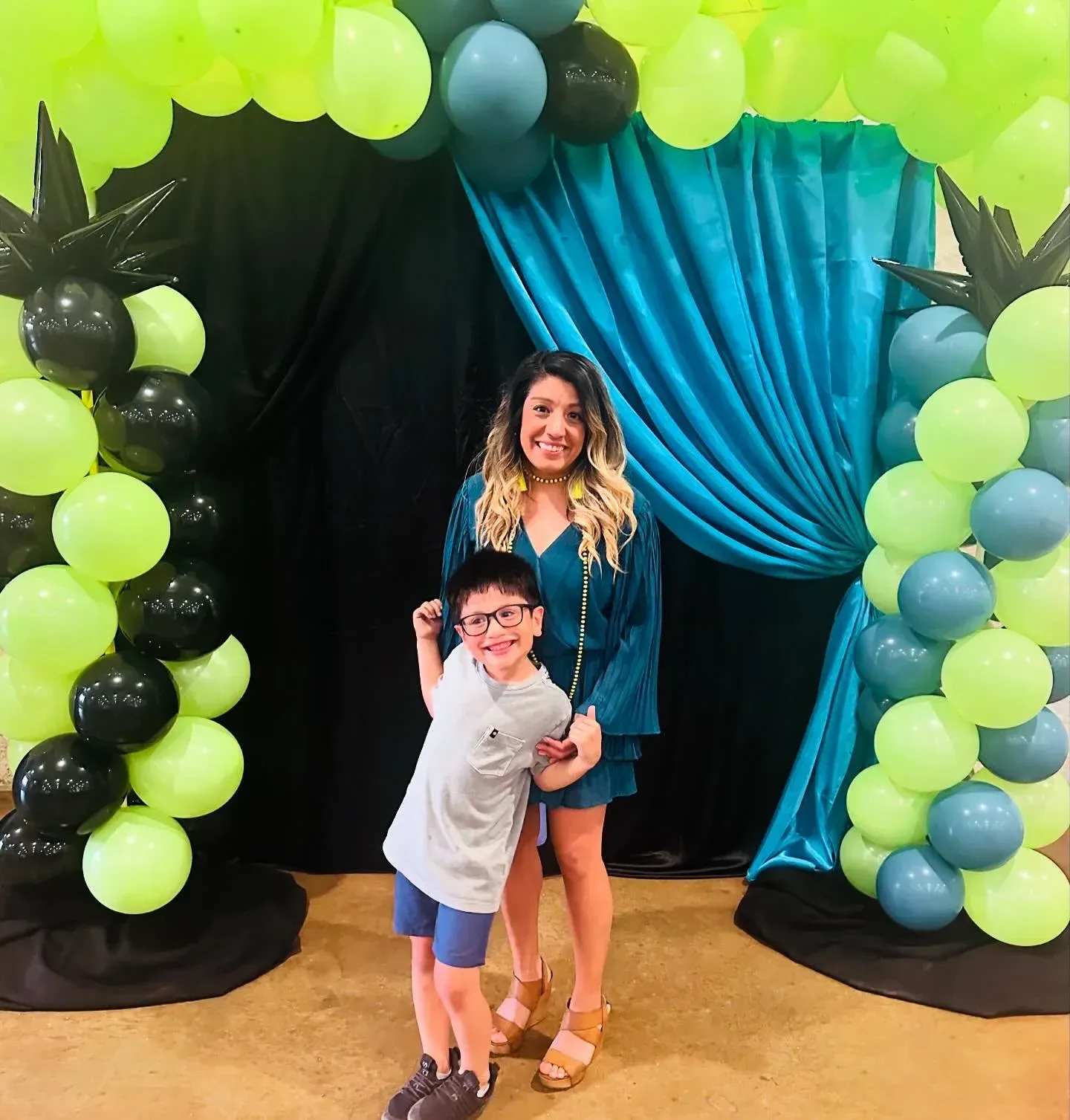 Woman and child pose under a balloon arch. Neon green, teal, and black balloons with teal backdrop.